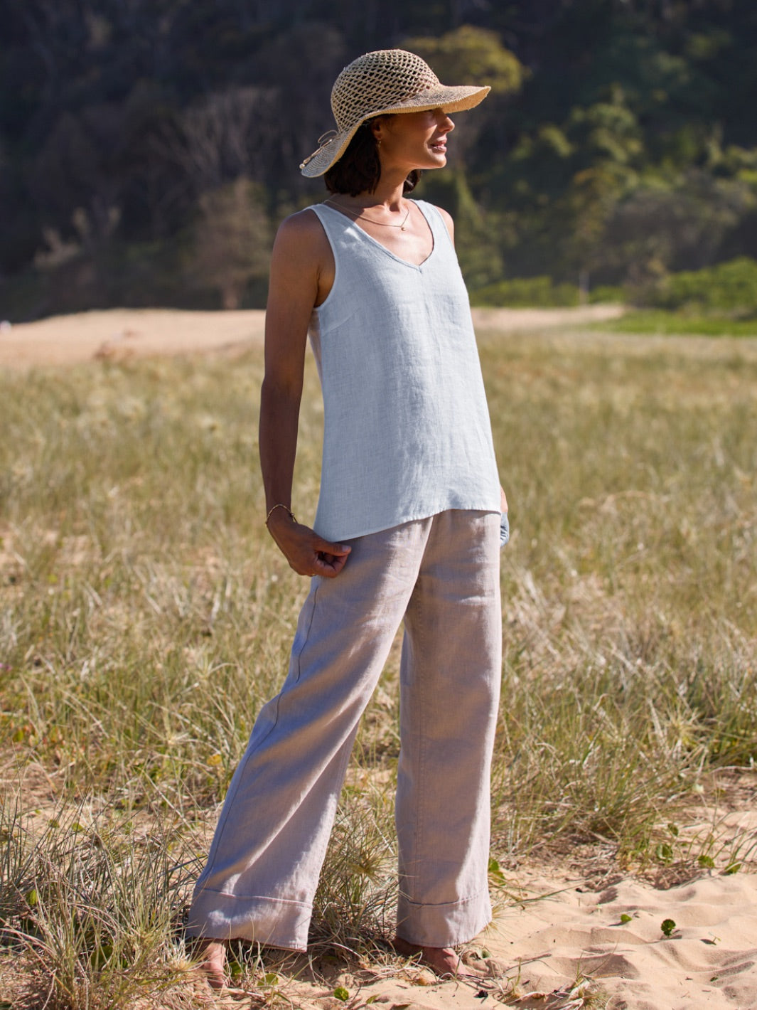 Woman wearing a light blue sleeveless top and beige pants, standing in a natural setting with trees and grass.