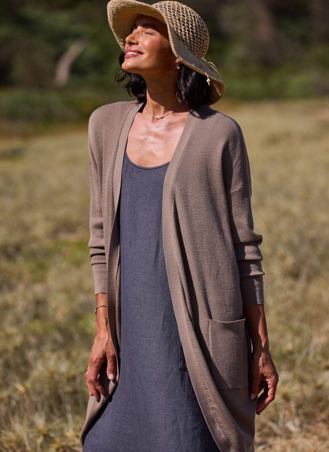 Woman wearing a long brown cardigan over a charcoal dress with a straw hat in a natural setting.