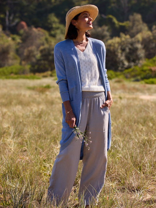 Woman in a blue cardigan and beige pants standing in a field with a straw hat on.