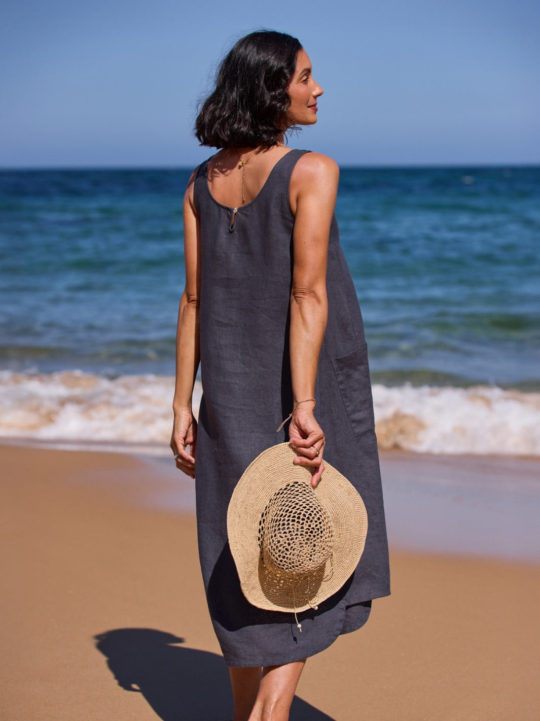 Woman in a dark dress holding a straw hat on a beach with ocean in the background
