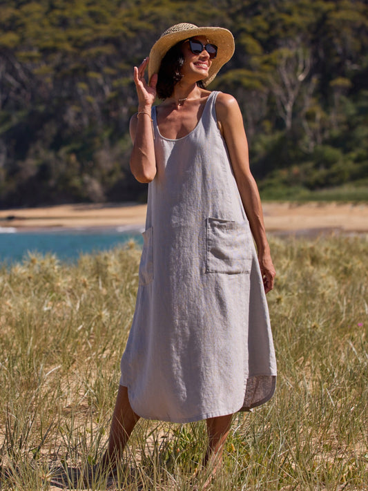 Woman in a gray dress and straw hat standing in a grassy field near a body of water.