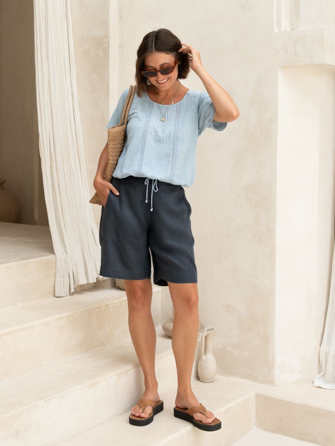 Woman wearing a light blue top and dark shorts, standing in a bright room.