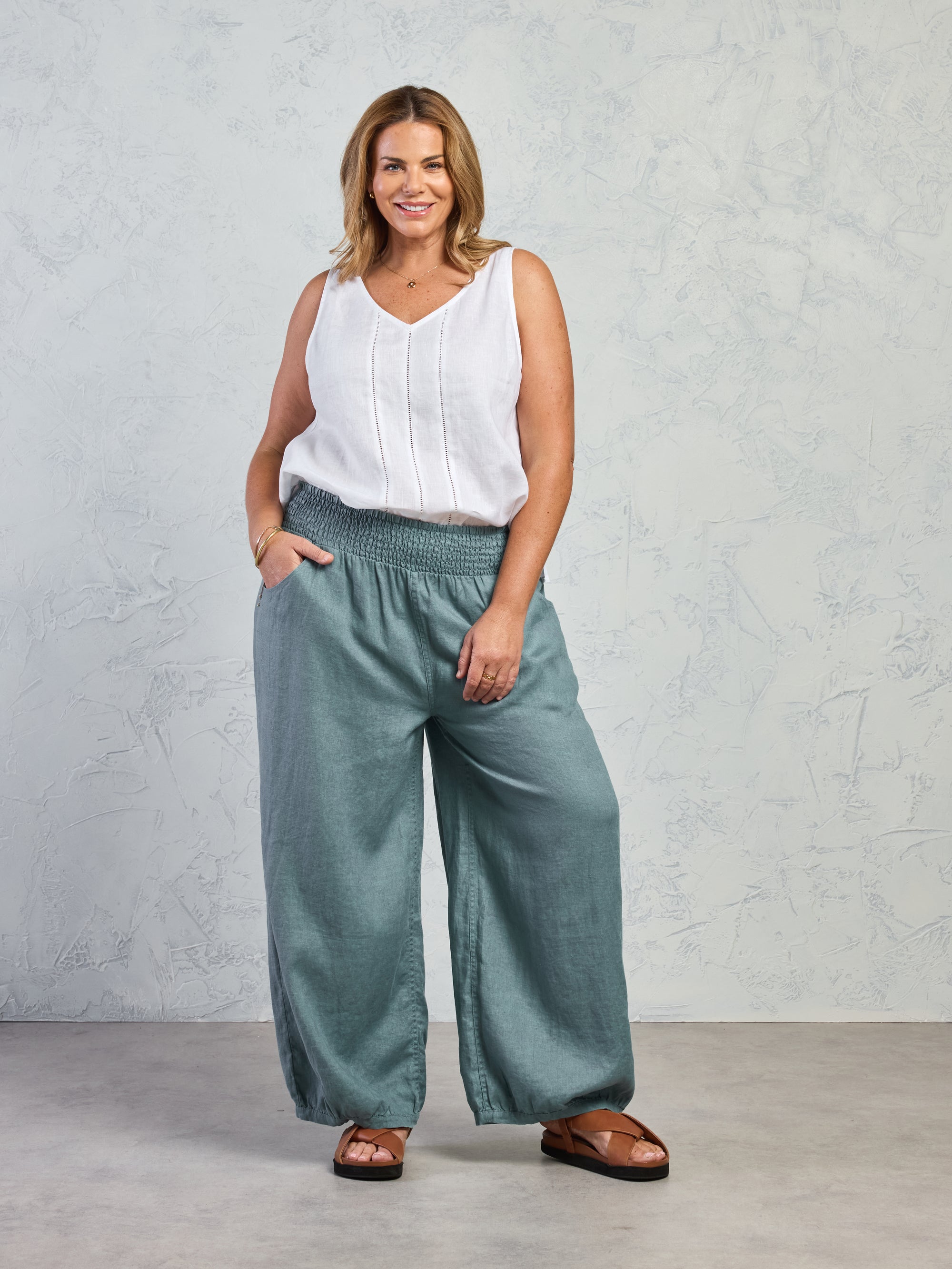 Woman wearing a white sleeveless top and teal wide-leg pants against a gray background