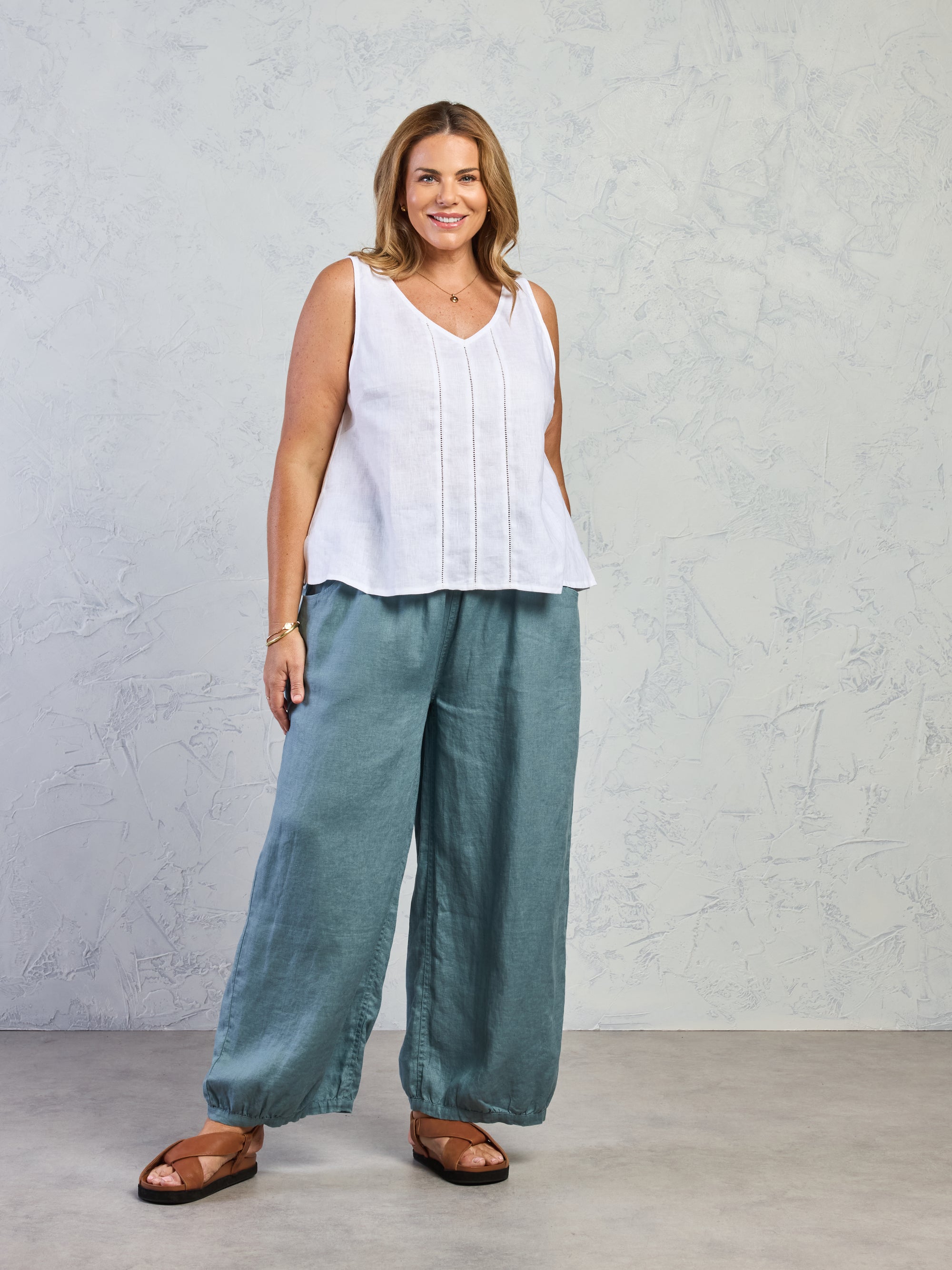 Woman wearing a white sleeveless top and teal pants against a plain background