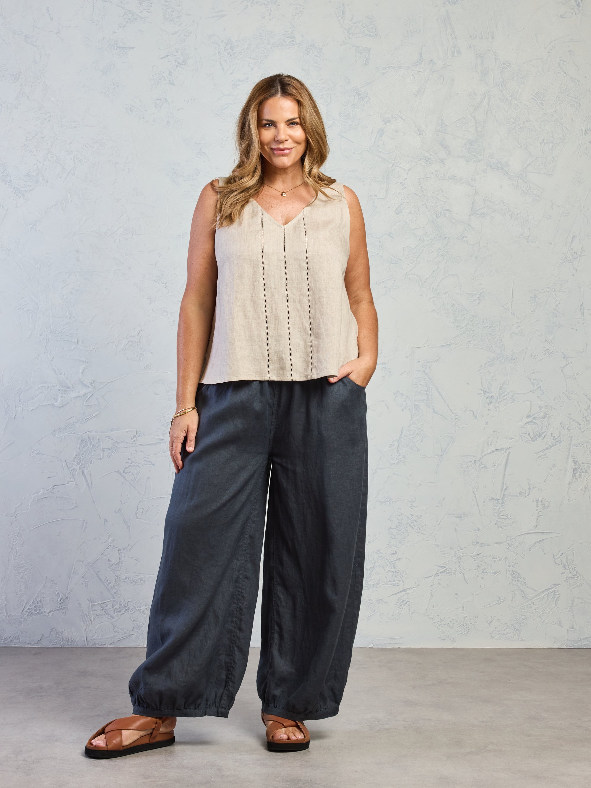 Woman wearing a beige sleeveless top and black pants against a gray background