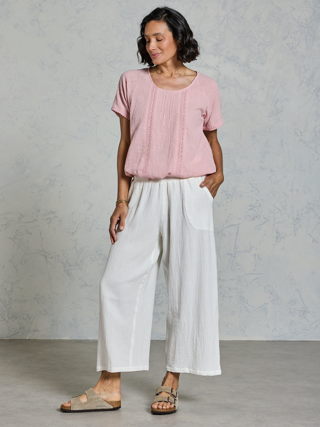 Woman wearing a pink top and white pants against a gray background