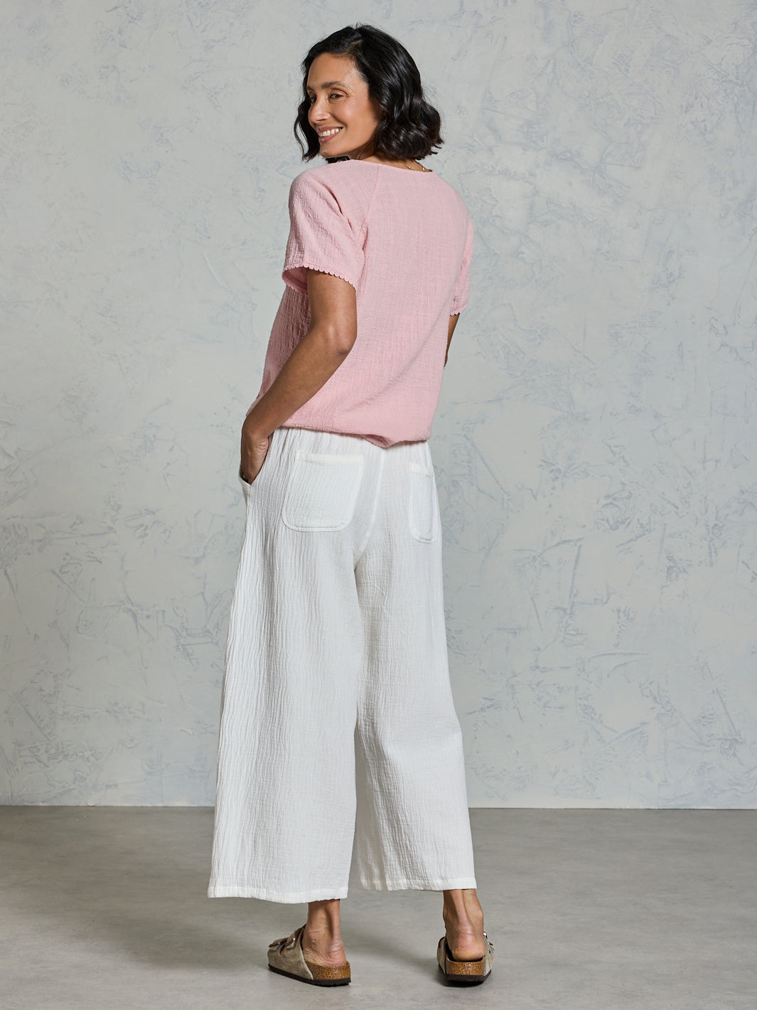 Woman wearing a pink top and white pants against a gray background