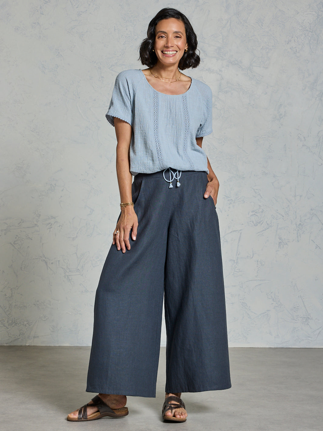 Woman wearing a light blue top and charcoal wide-leg pants against a plain background