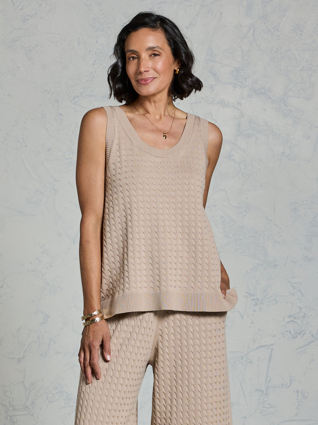 Woman wearing a beige knitted sleeveless top and pants against a light gray background