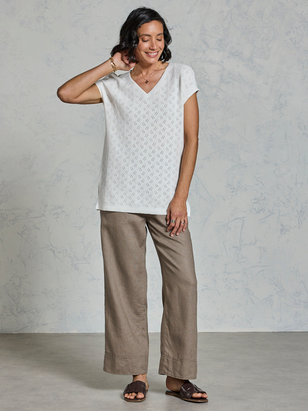 Woman wearing a white patterned top and beige pants against a gray background