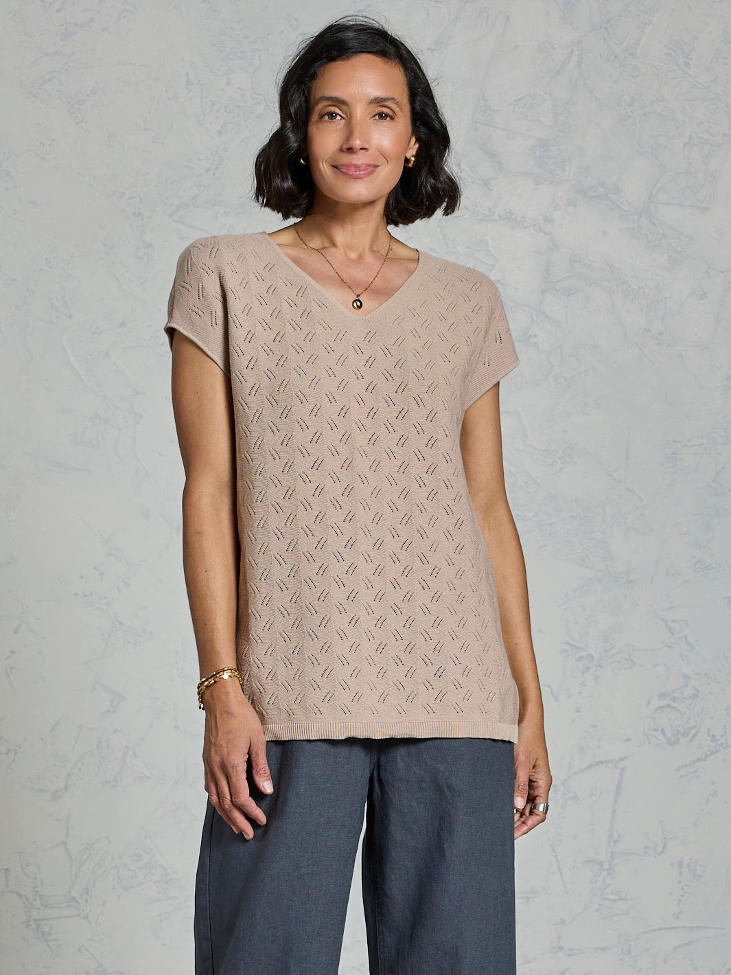 Woman wearing a beige patterned top and dark pants against a light gray background