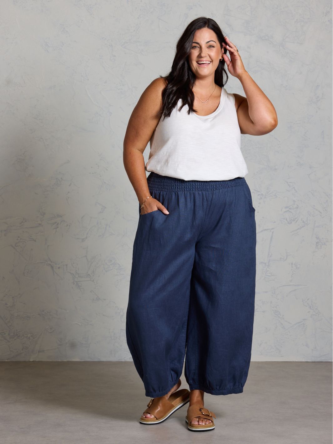 Woman wearing a white tank top and dark pants against a gray background