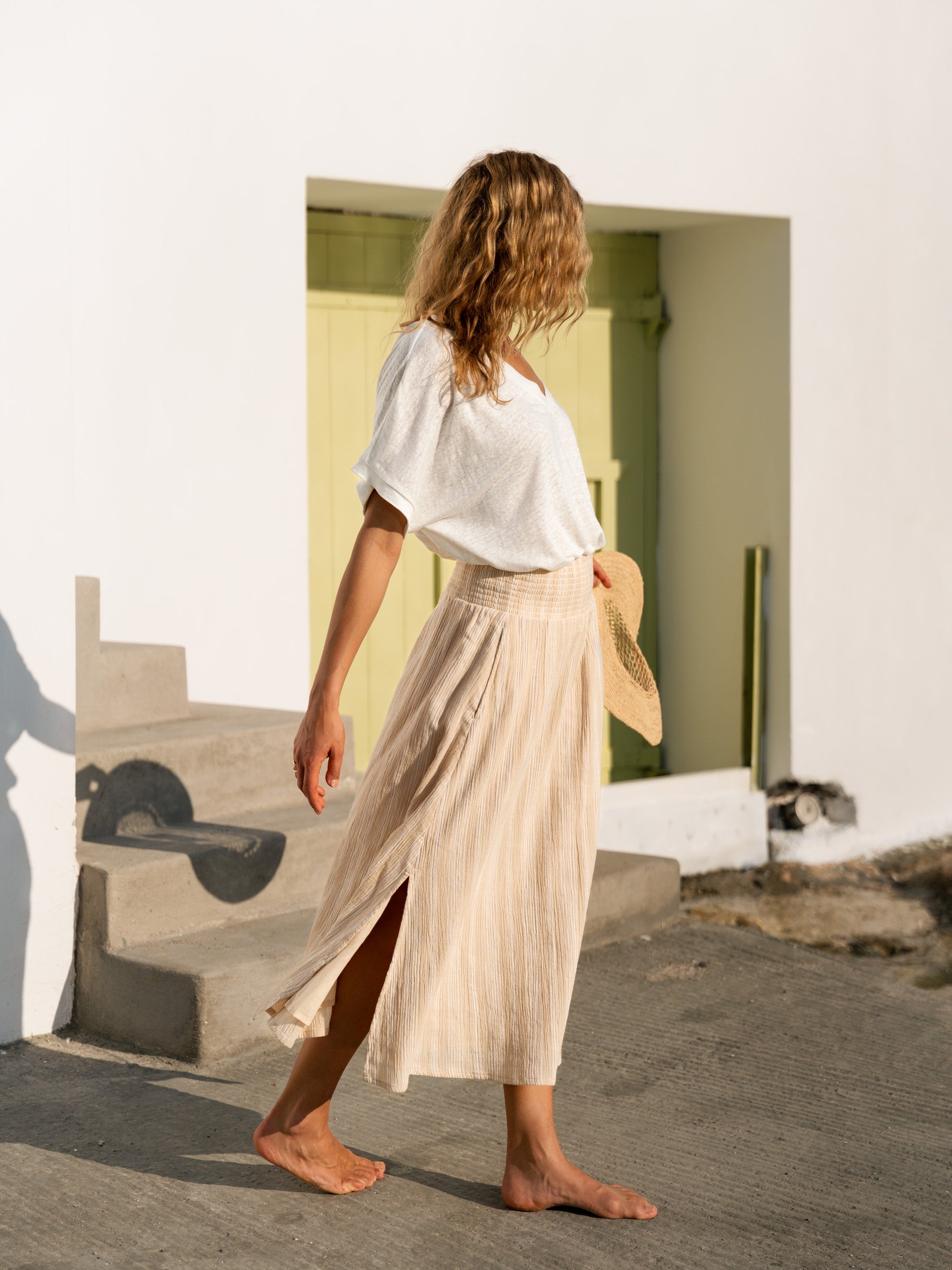 Woman in a white linen top and beige cotton skirt holding a woven hat