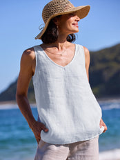 Woman wearing a light blue sleeveless top and beige hat on a beach with mountains in the background