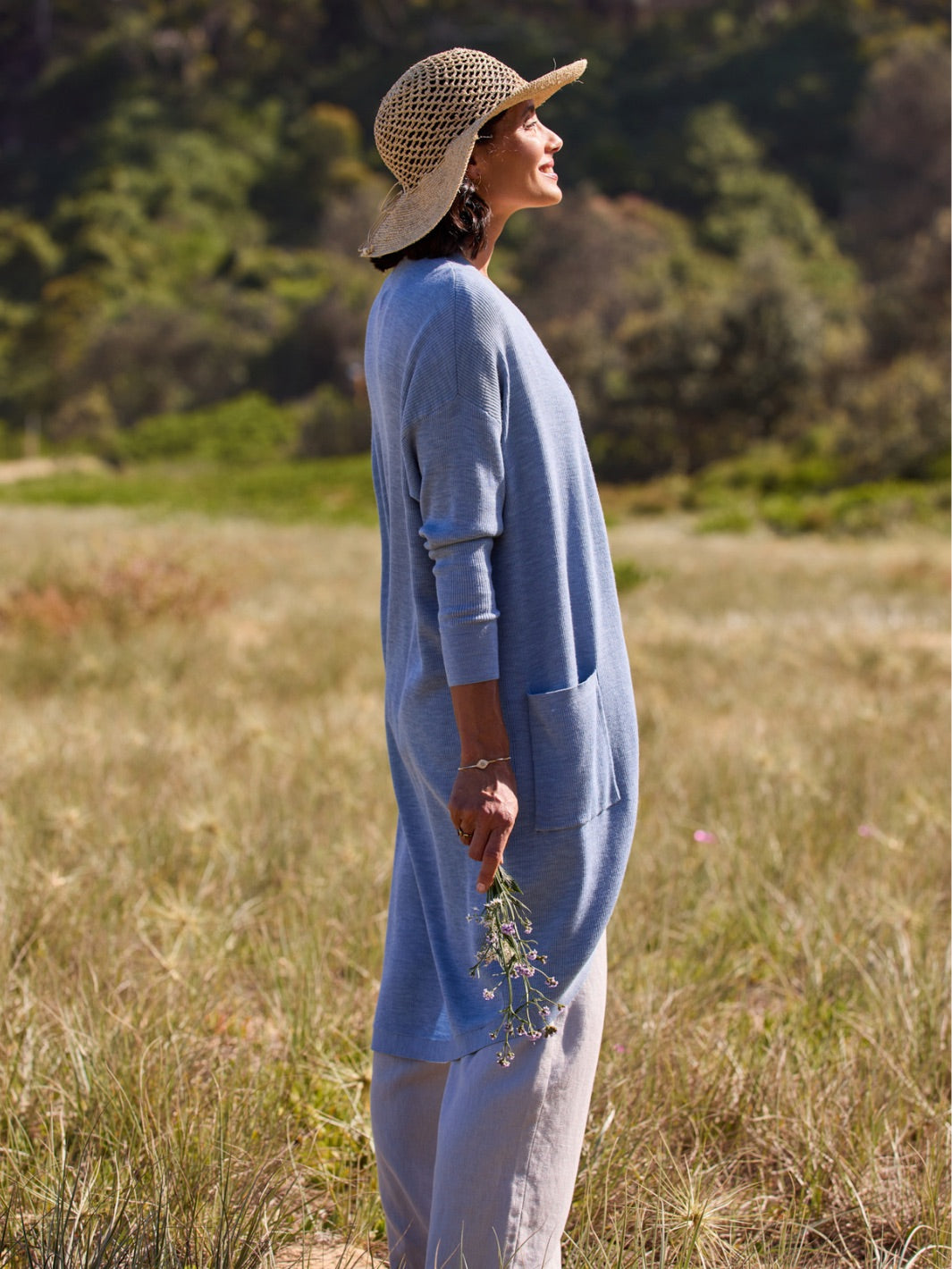 Woman in a blue dress and straw hat standing in a field with trees in the background