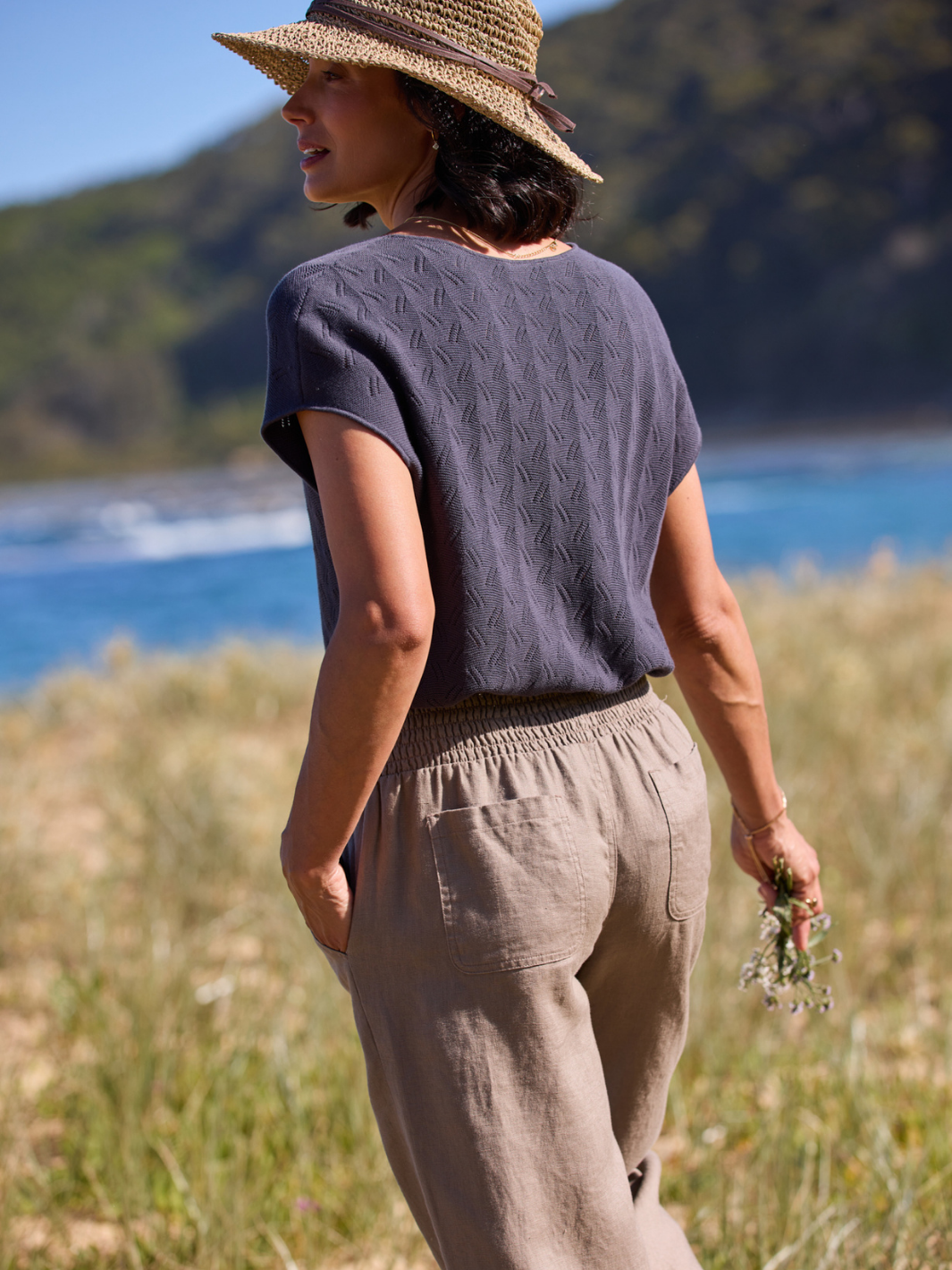 Person wearing a dark knitted top and beige pants standing in a natural setting with mountains and water in the background.
