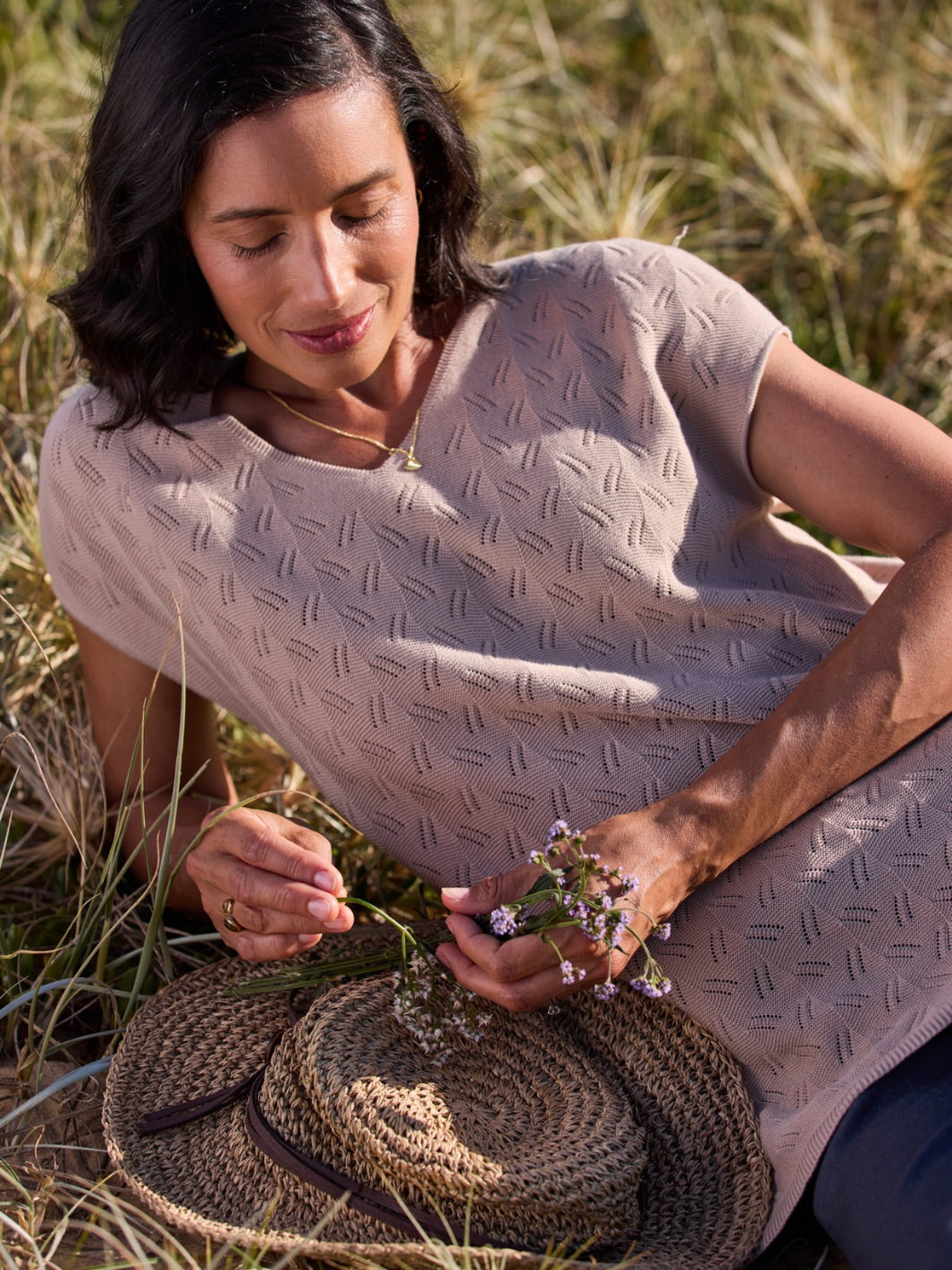 Woman sitting in a field holding a woven basket and flowers, wearing a light-colored patterned top.