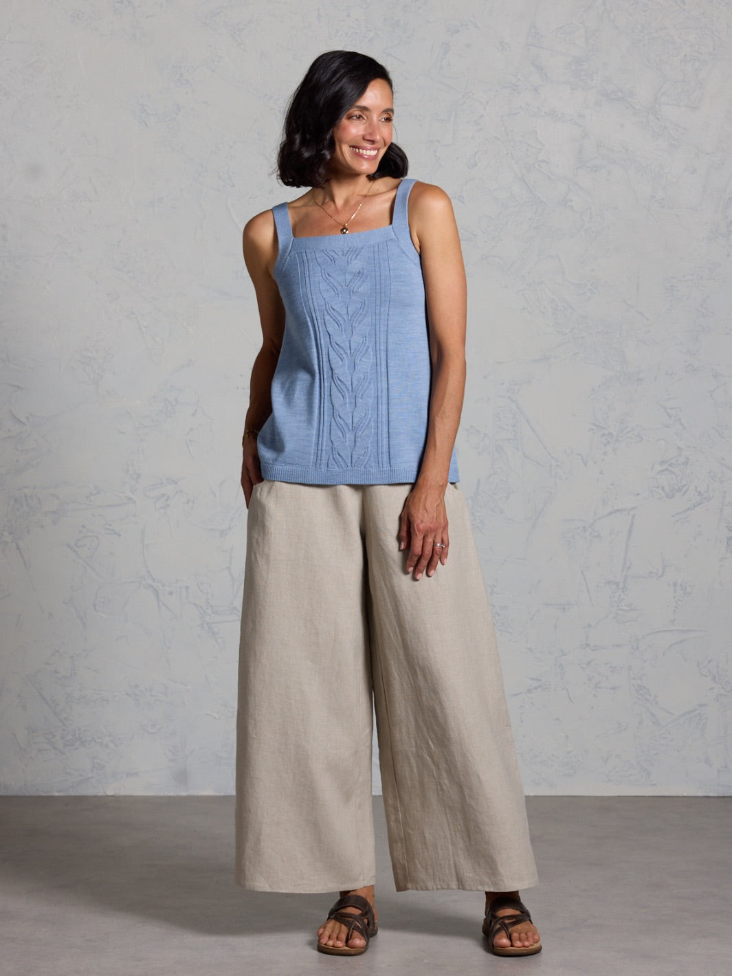 Woman wearing a blue sleeveless top and beige wide-leg pants against a gray background