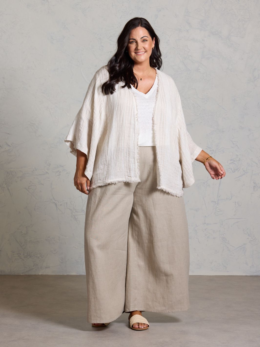 Woman wearing a white top and beige pants with a white shawl against a plain background