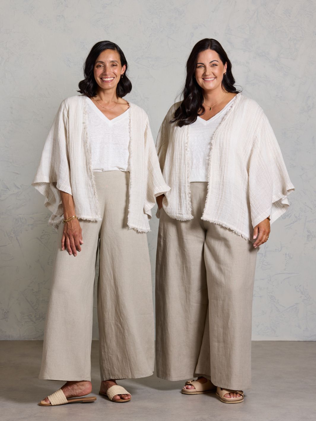 Two women wearing matching outfits with white tops and beige pants against a plain background