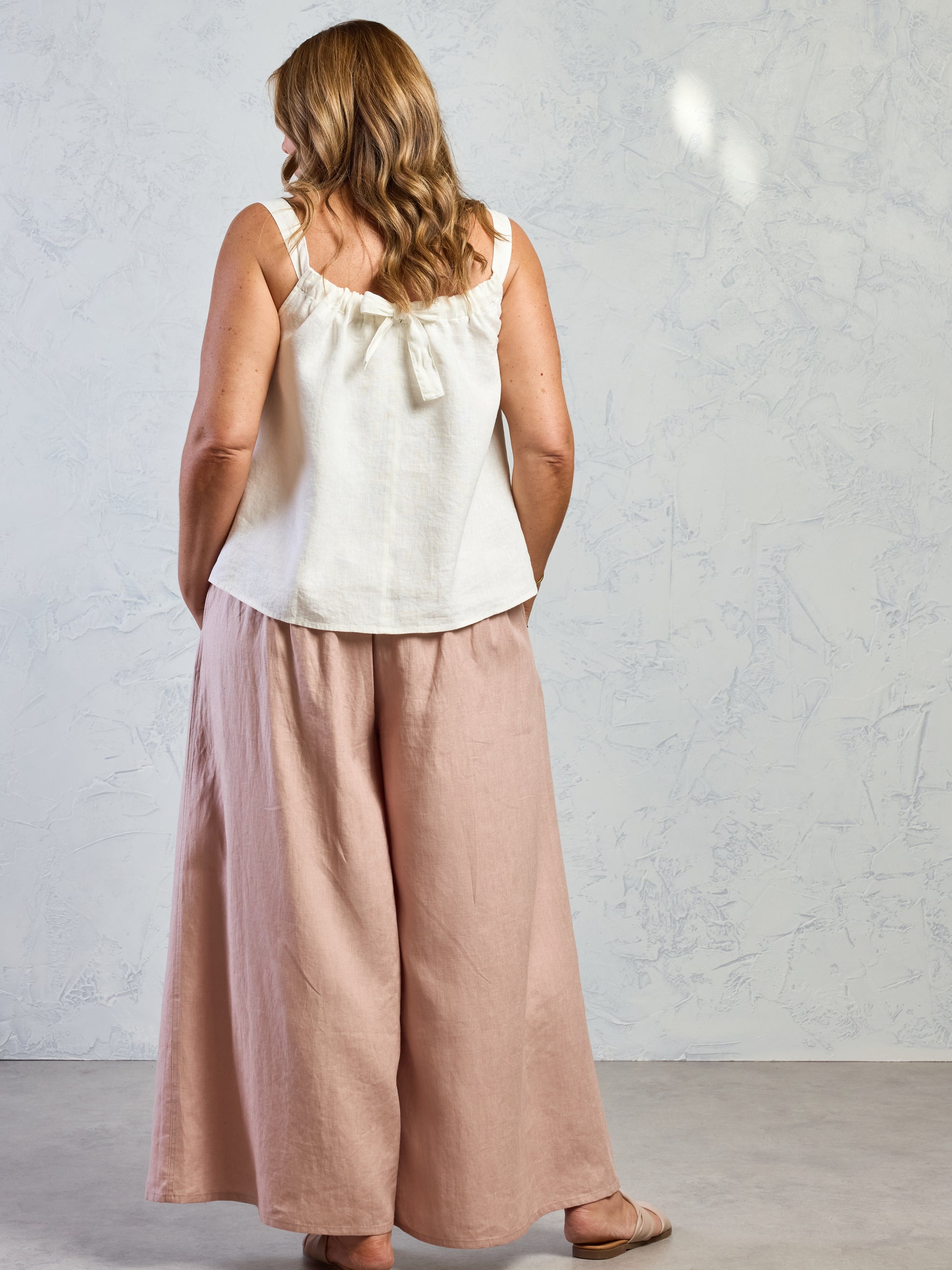 Woman wearing a white sleeveless top and pink wide-leg pants against a gray background