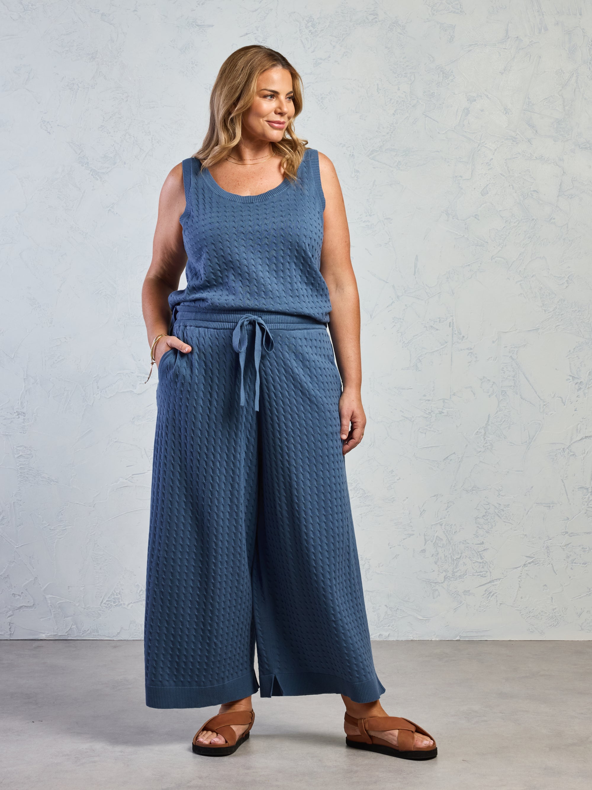 Woman wearing a blue textured sleeveless top and wide-leg pants against a light gray background