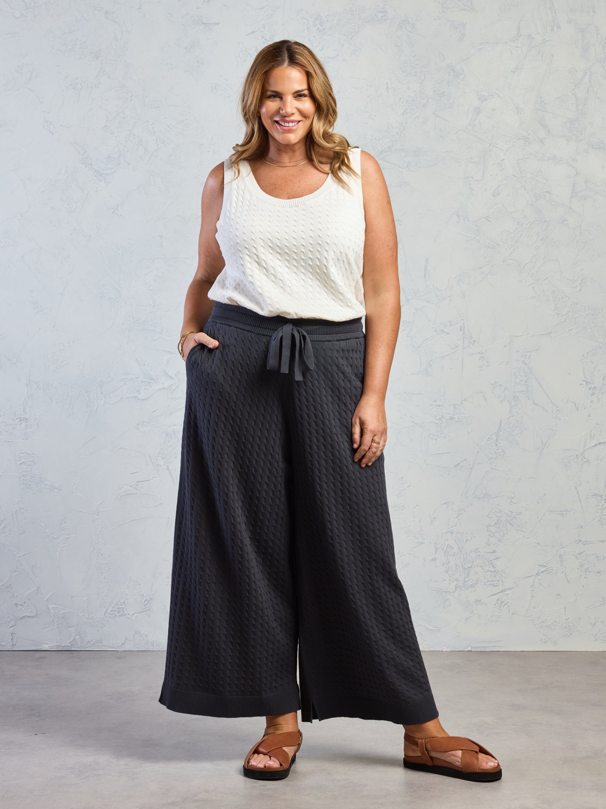 Woman wearing a white sleeveless top and dark wide-leg pants against a gray background