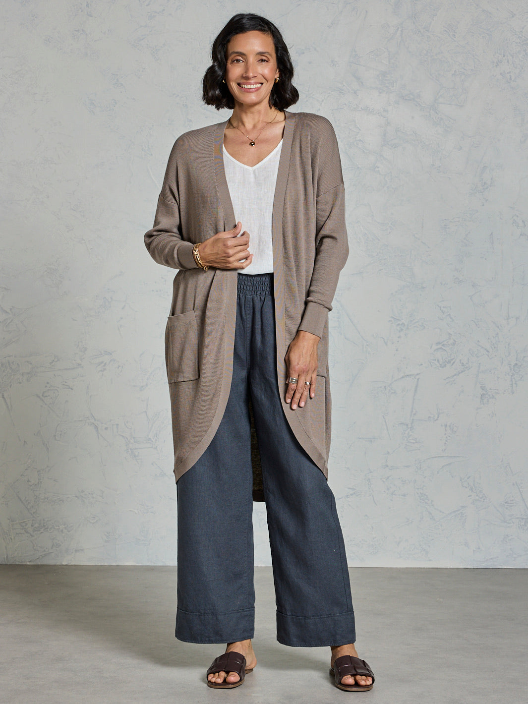 Woman wearing a long beige cardigan over a white top and dark blue pants against a plain background