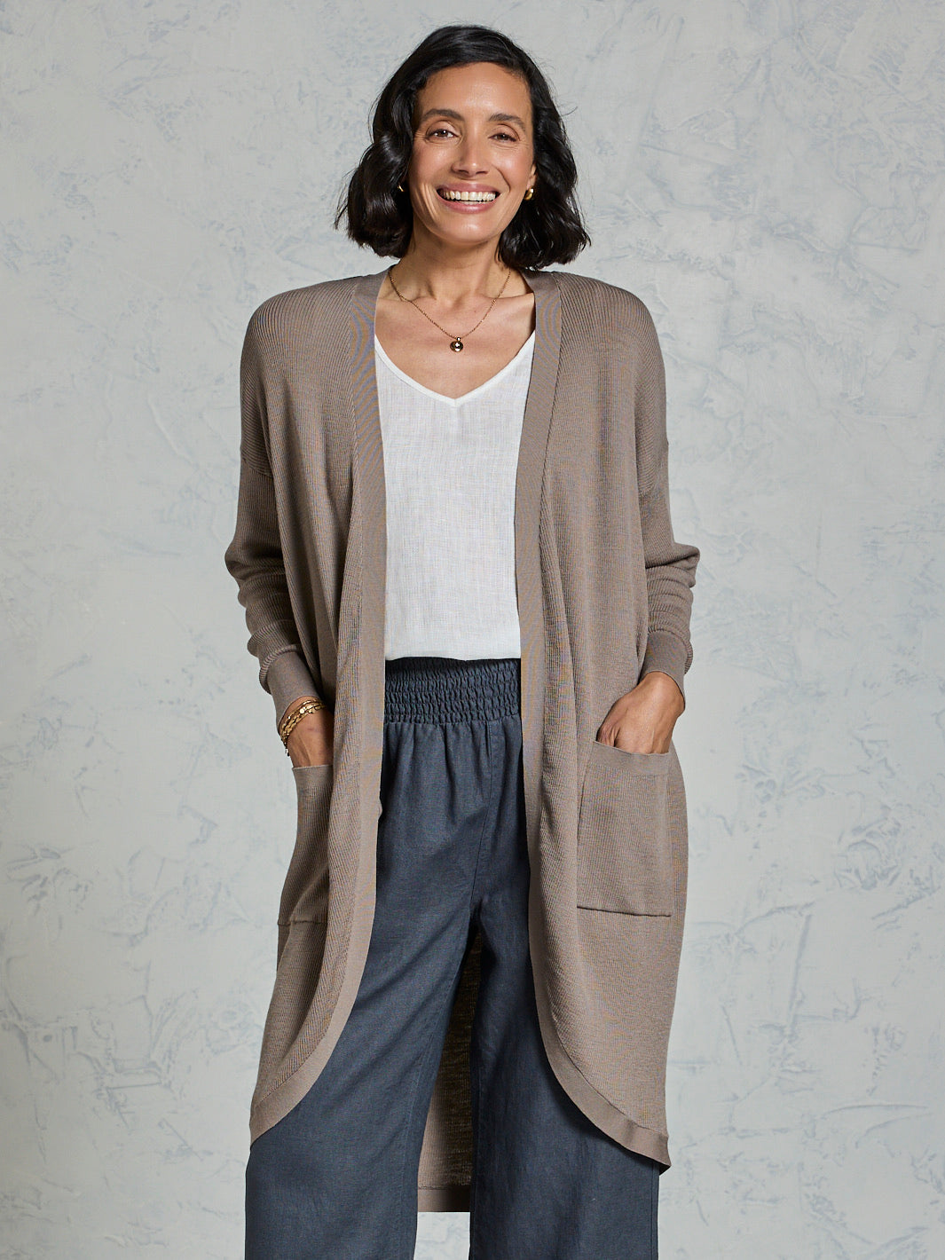 Woman wearing a long beige cardigan over a white top and dark pants on a light gray background