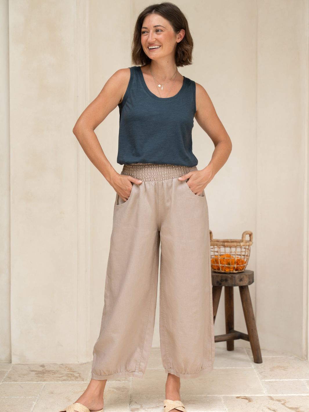 Woman wearing a black tank top and beige wide-leg pants standing in a room with a wooden stool and basket in the background.