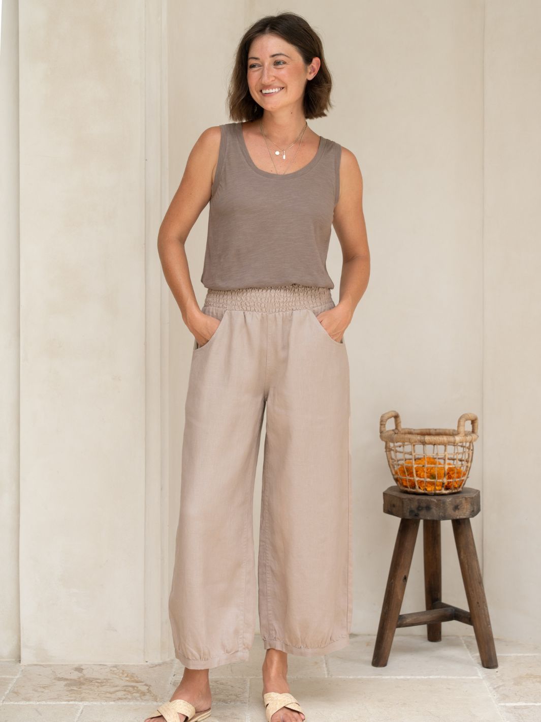 Woman wearing a sleeveless top and wide-leg pants standing next to a wooden stool with a basket.