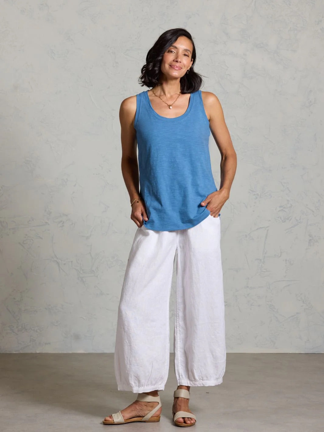 Woman wearing a blue tank top and white pants on a grey background.