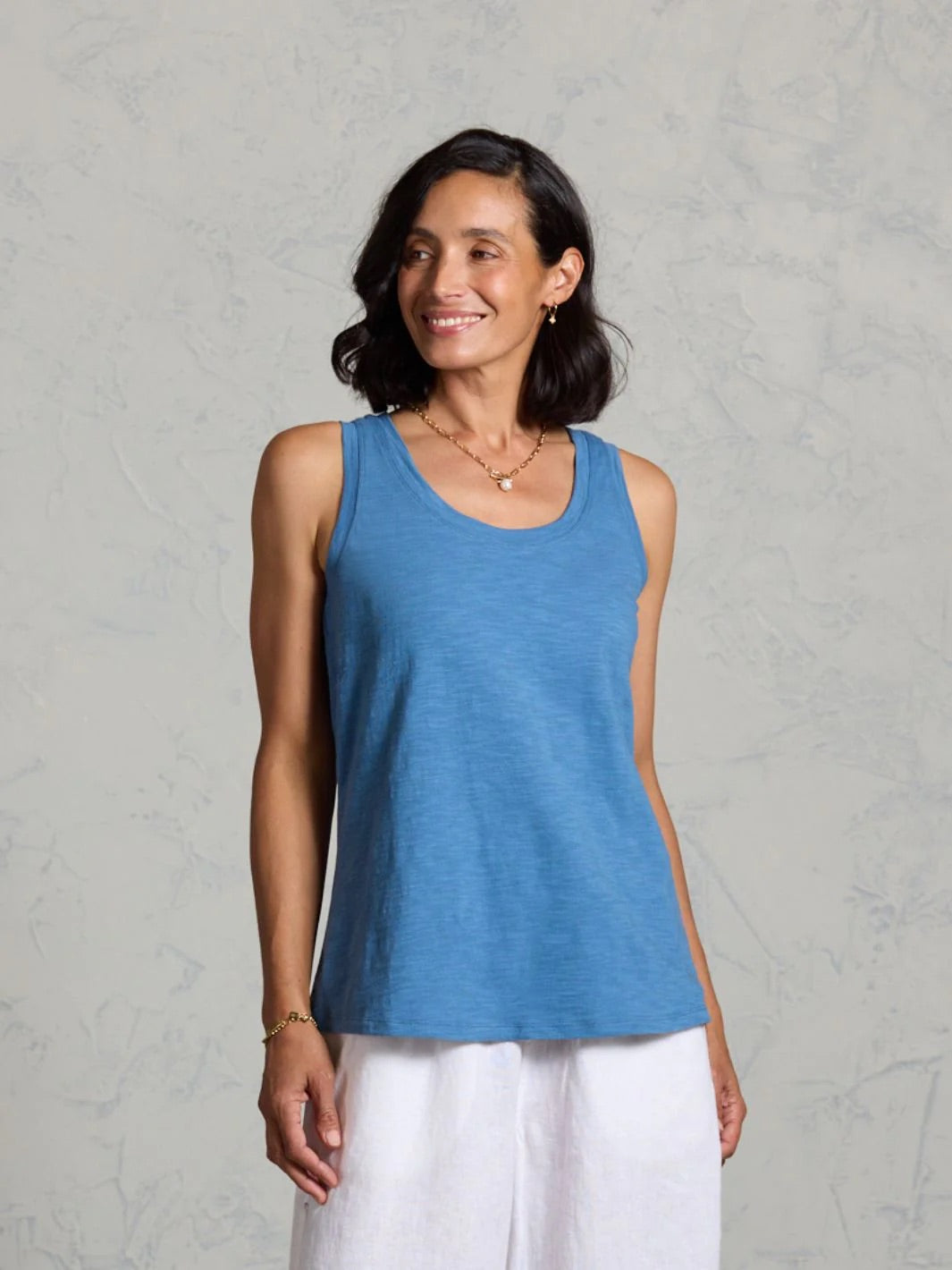 Woman wearing a blue tank top and white pants on a grey background. 