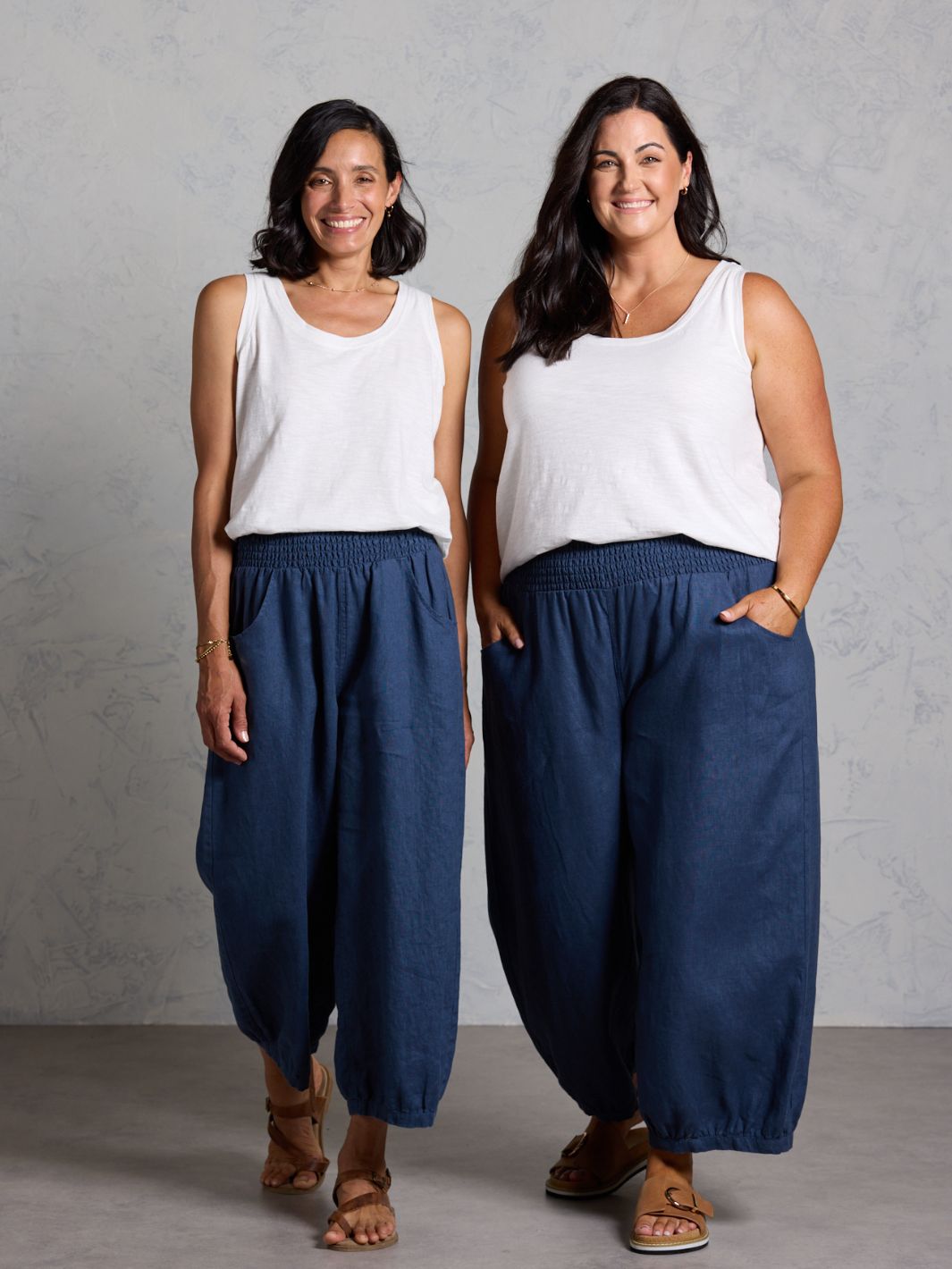 Two women wearing white tops and dark pants standing against a gray background
