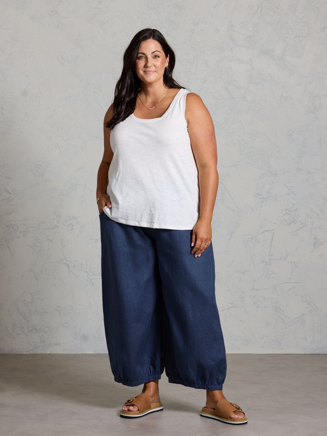 Woman wearing a white sleeveless top and dark pants against a gray background