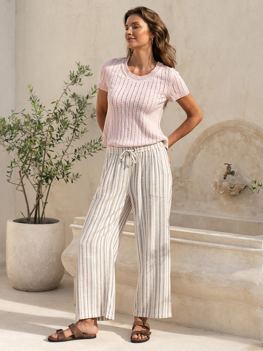 Model wearing the Jolie Linen Blend Pants in driftwood with a pink knit top, beside an olive tree in a sunlit Mediterranean courtyard.